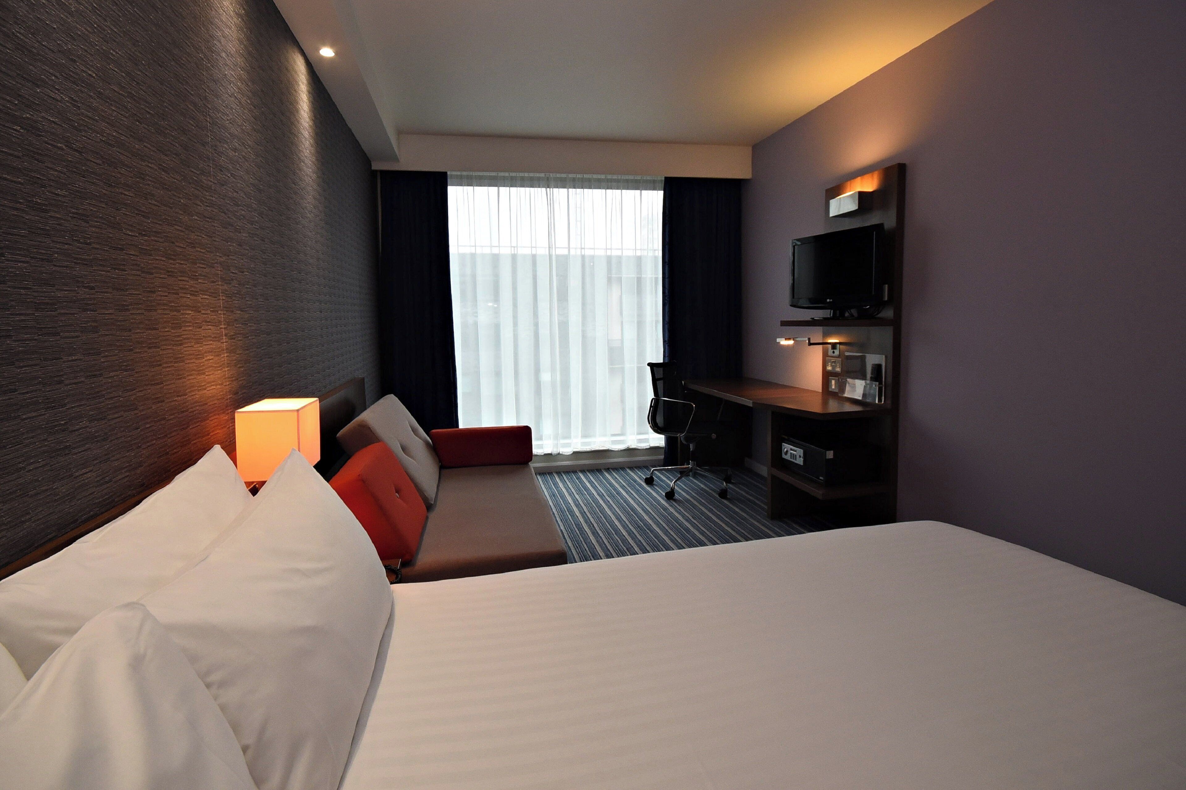 A hotel room with a bed, a desk, a chair, a TV, and a window with curtains.