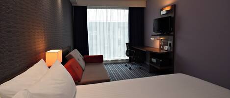 A hotel room with a bed, a desk, a chair, a TV, and a window with curtains.