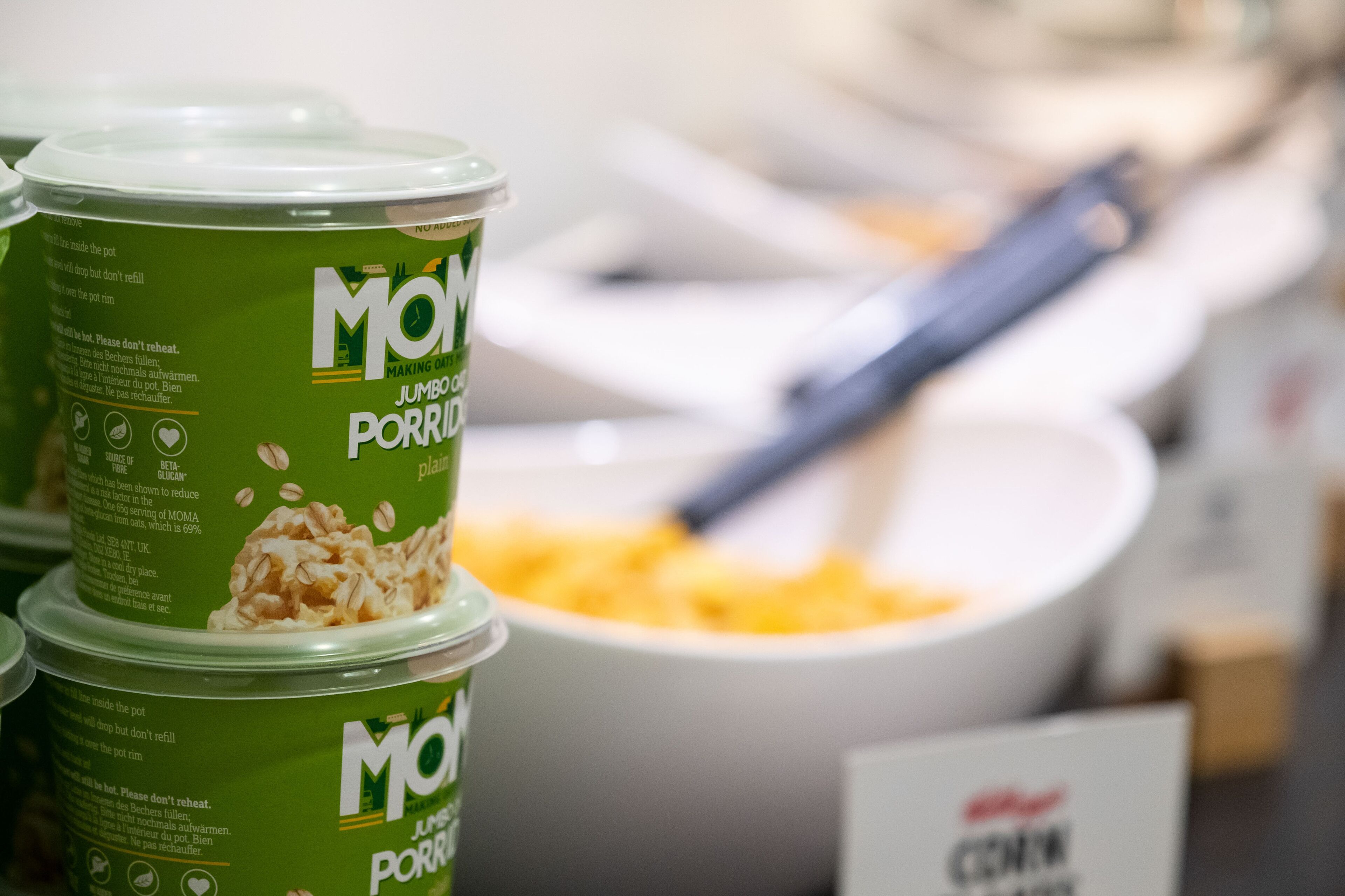 A container of MOMA jumbo porridge with a spoon in a bowl of porridge in the background.