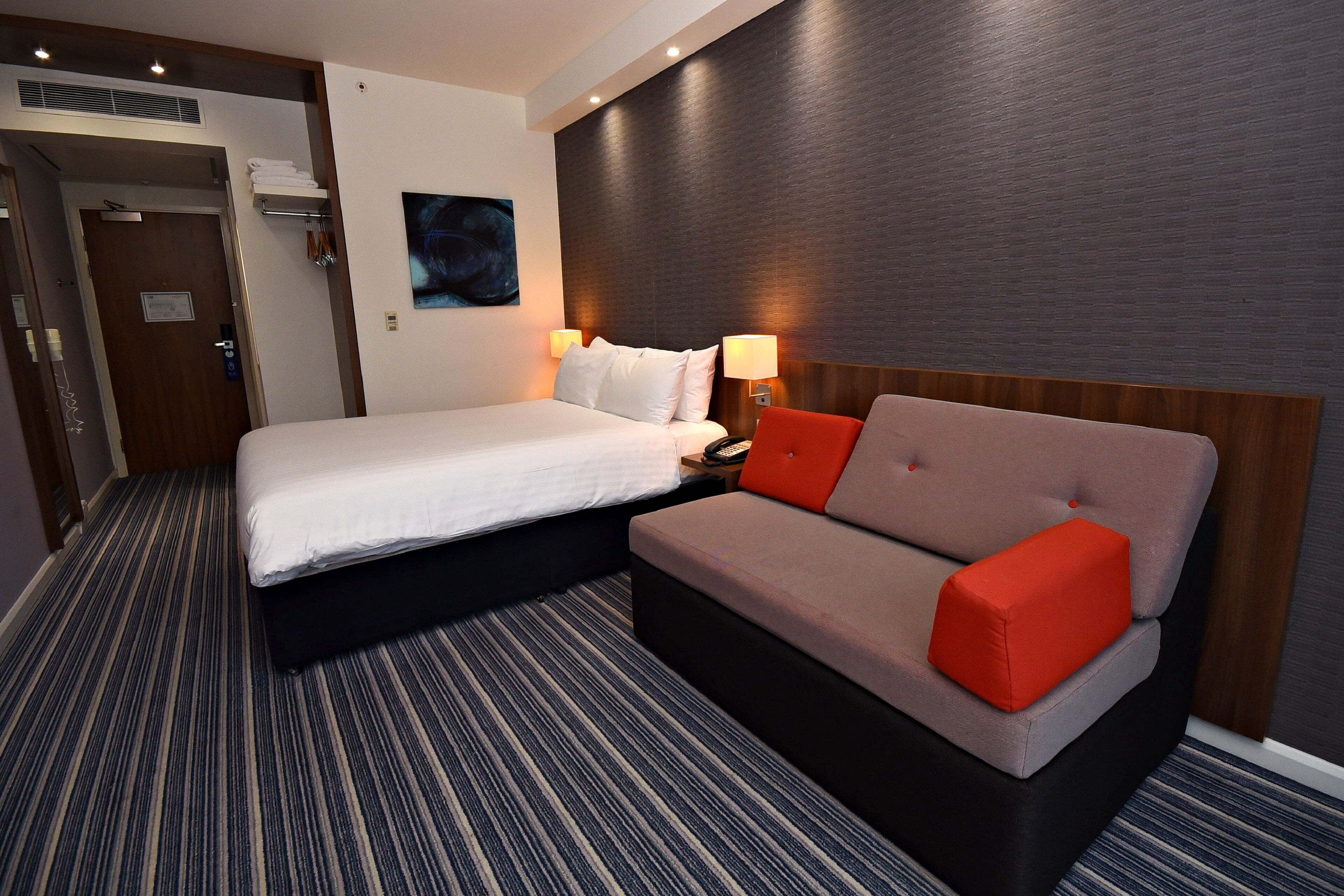 A hotel room with a bed, a sofa, a nightstand, and a wall-mounted lamp.