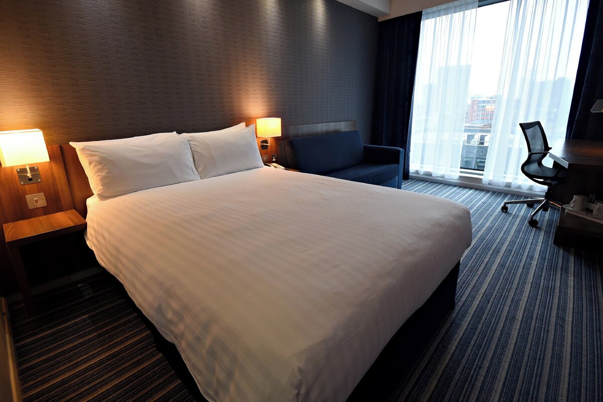 A hotel room with a large bed, two bedside tables with lamps, a blue armchair, a desk with a chair, and a large window with curtains.