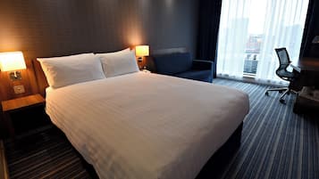 A hotel room with a large bed, two bedside tables with lamps, a blue armchair, a desk with a chair, and a large window with curtains.