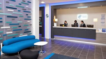 Un área de recepción de hotel con diseño moderno, que incluye un mostrador de recepción, un sofá azul y una pequeña mesa redonda.