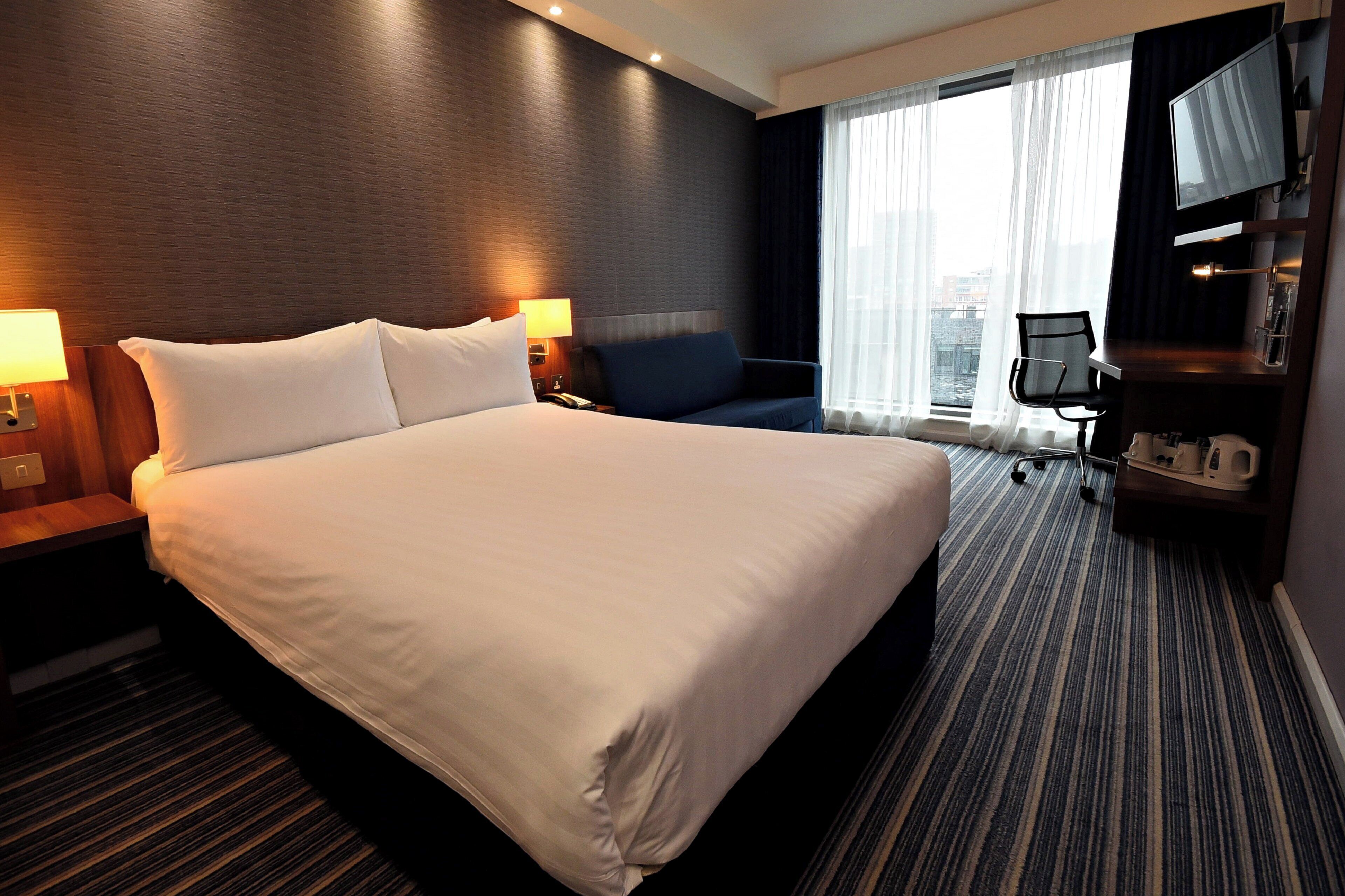 A hotel room with a large bed, two bedside tables with lamps, a sofa, a desk with a chair, a television, and a view of the city through the window.