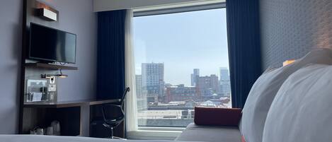 A hotel room with a bed, a desk with a flat-screen TV, a chair, and a window with a city view.