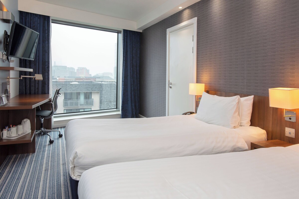 A hotel room with two beds, a desk with a chair, a TV mounted on the wall, and a window with a view of the city.