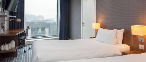 A hotel room with two beds, a desk with a chair, a TV mounted on the wall, and a window with a view of the city.
