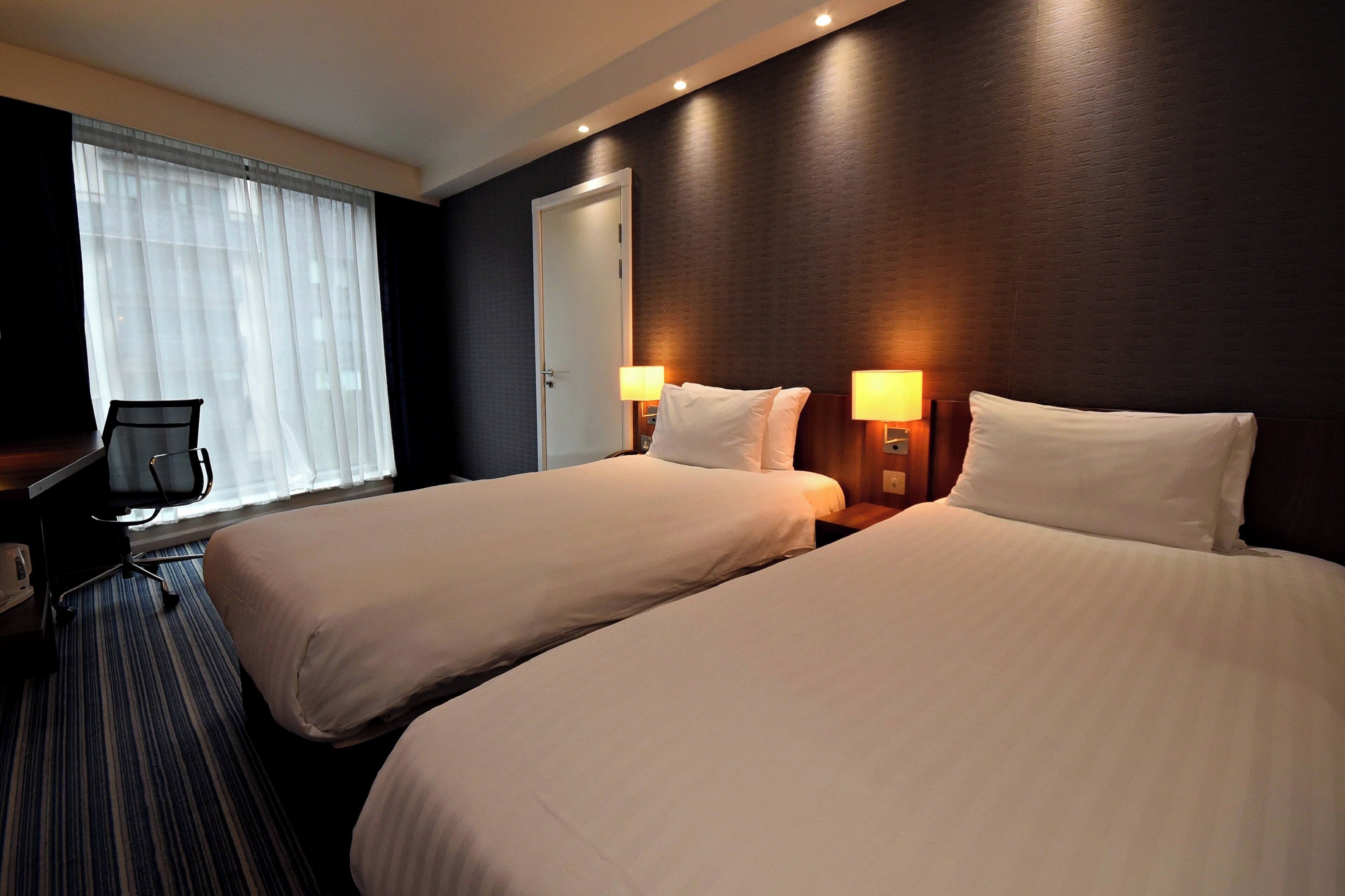 A hotel room with two beds, a desk, a chair, and a window with sheer curtains.