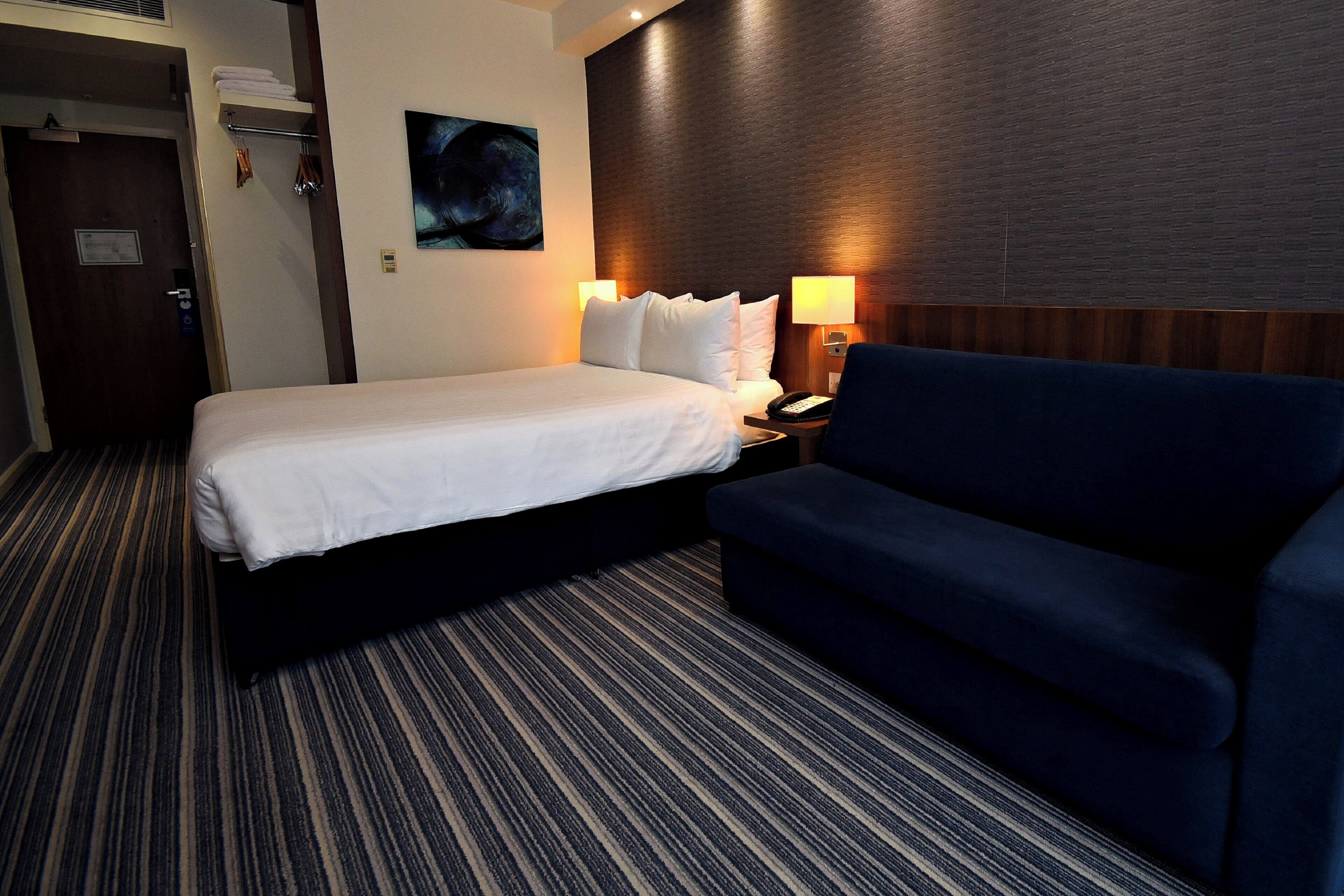 Una habitació d'hotel amb un llit, un sofà blau, una taula de nit amb una làmpada, un quadre a la paret i una porta amb un assecador de tovalloles.