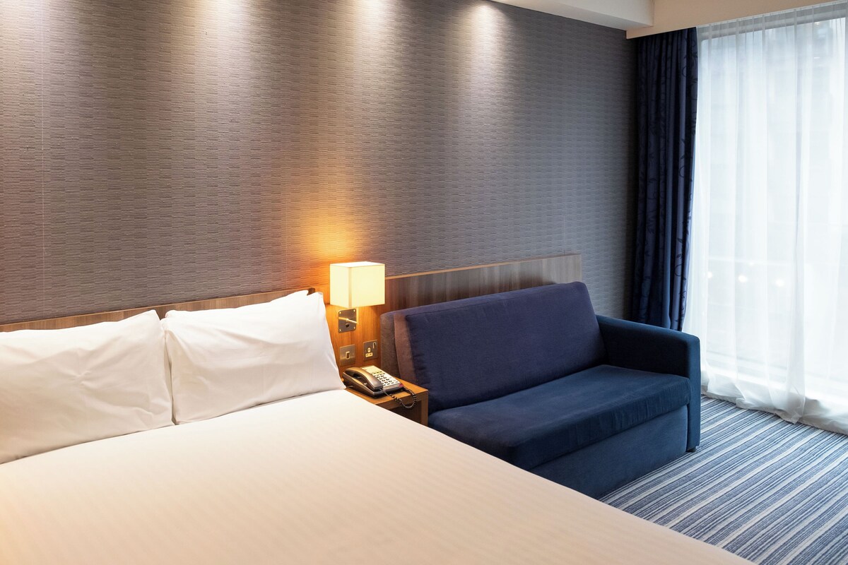 A hotel room with a bed, a nightstand with a lamp, a blue sofa, and a large window with curtains.