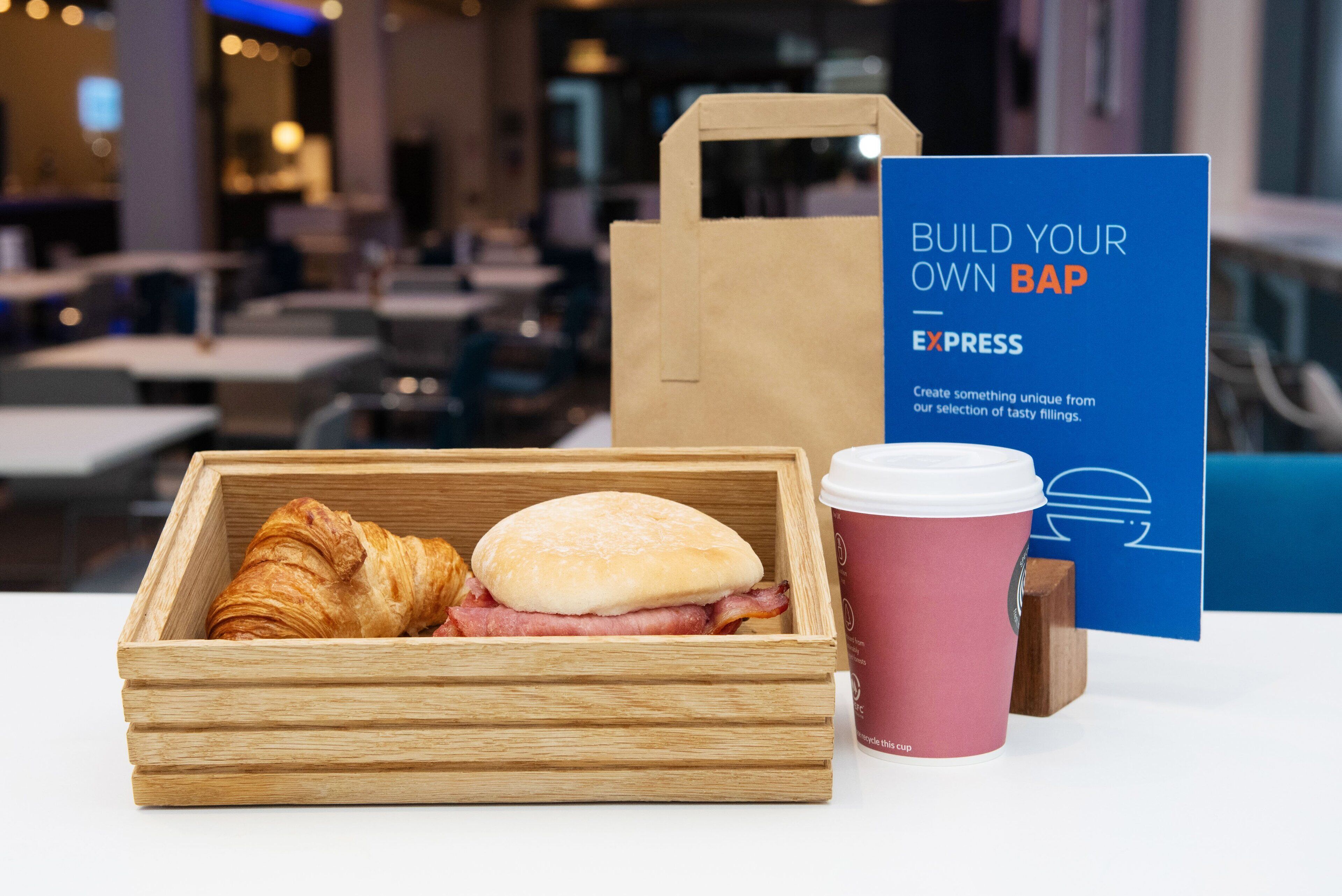 A wooden box with a croissant and a sandwich, a pink coffee cup, and a blue sign with text.