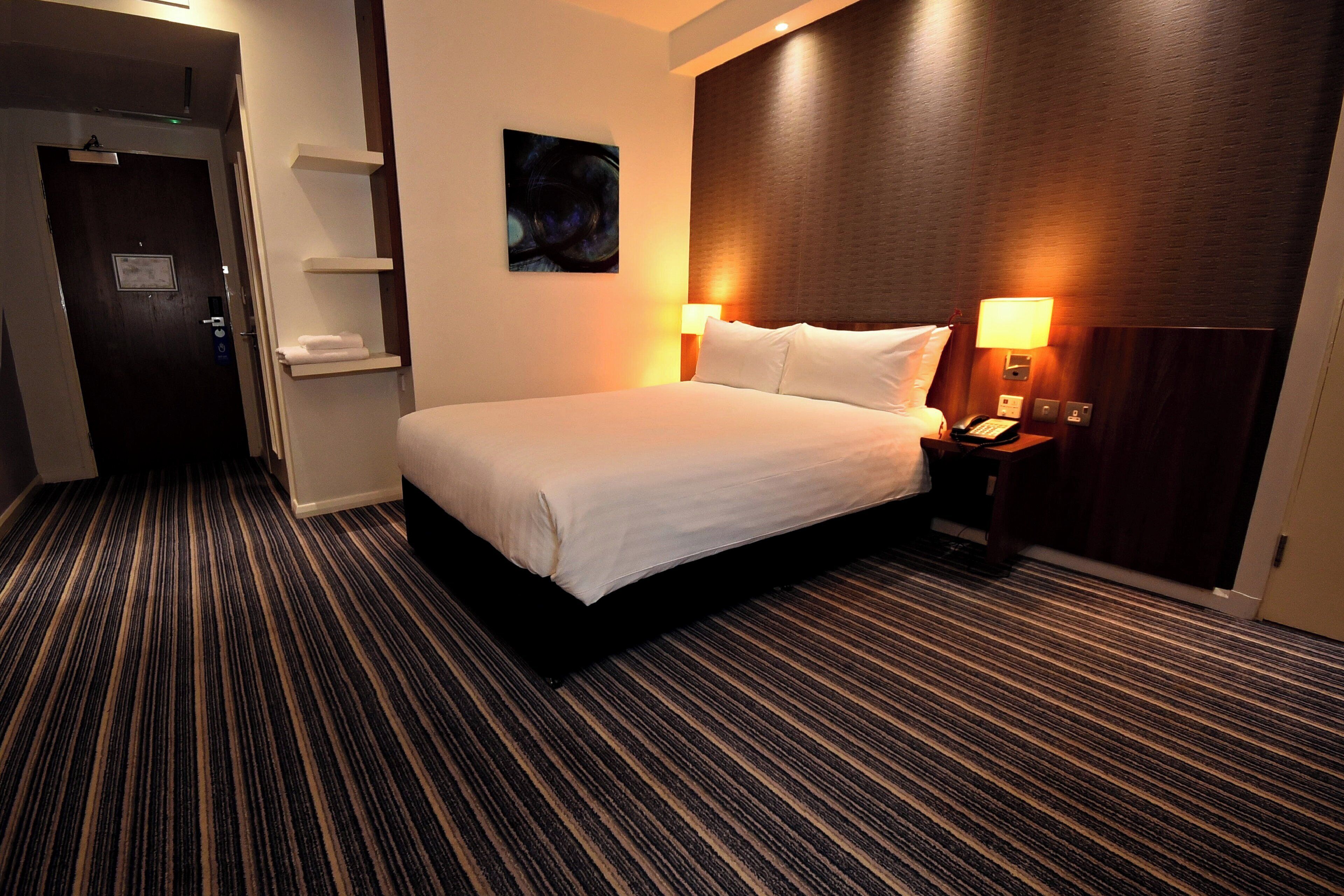 A hotel room with a bed, two bedside lamps, a nightstand, a wall-mounted artwork, and a door with a towel rack.