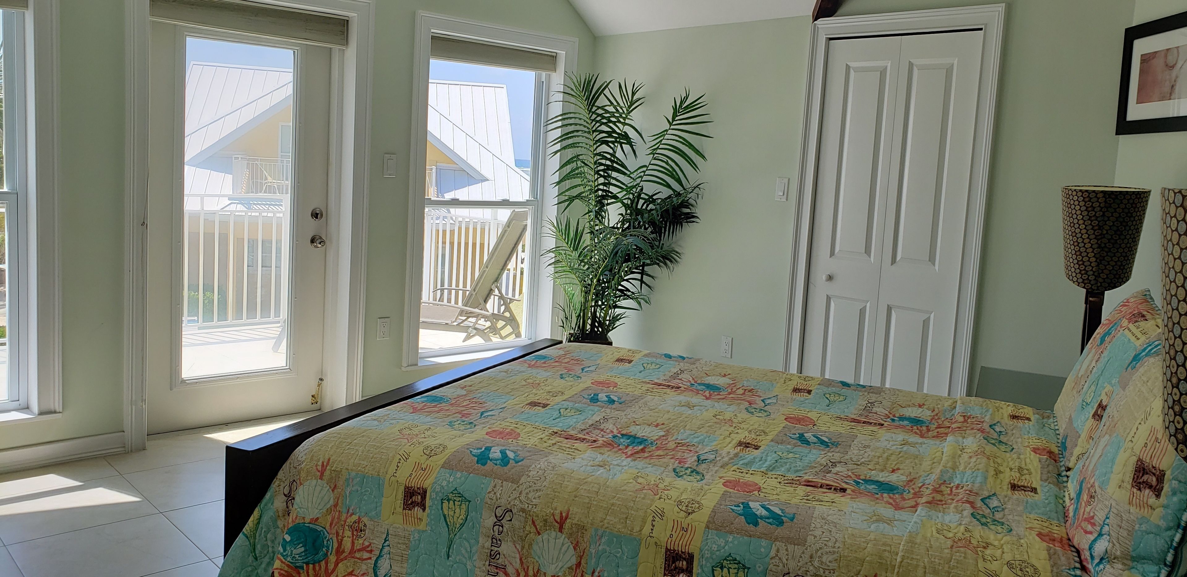 A bedroom with a large window, a bed with a colorful quilt, a potted plant, and a door.