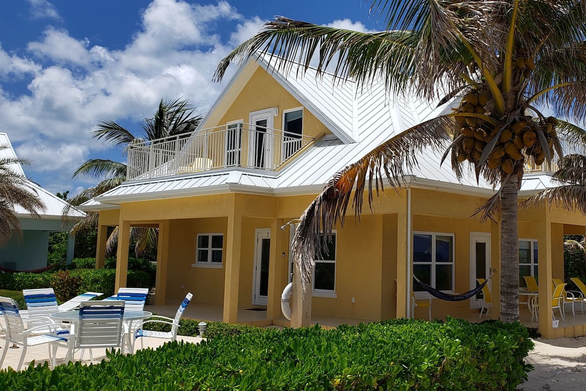 A yellow two-story house with a balcony, surrounded by palm trees and a sandy beach.