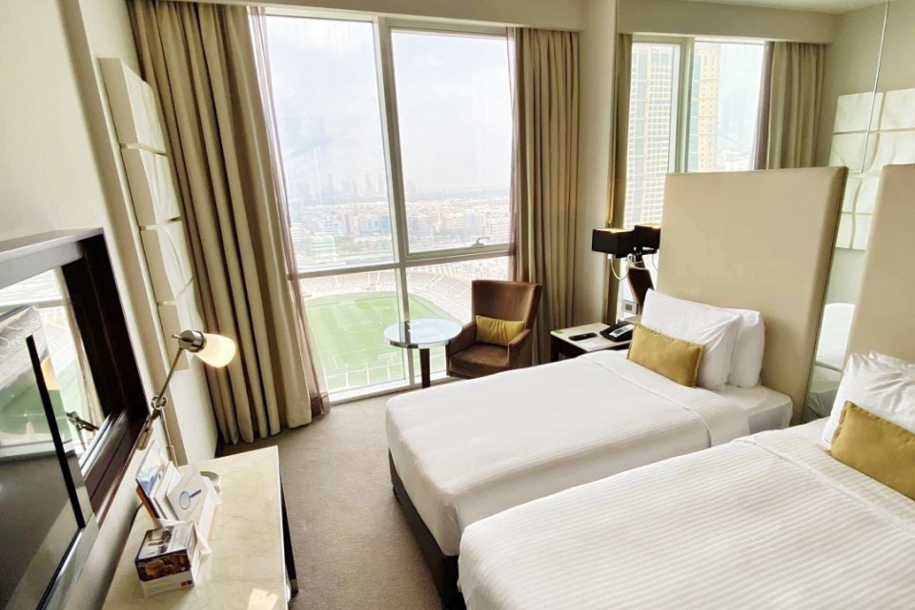 Habitación de hotel con dos camas, un escritorio, una silla, un televisor y un amplio ventanal con vistas a la ciudad.
