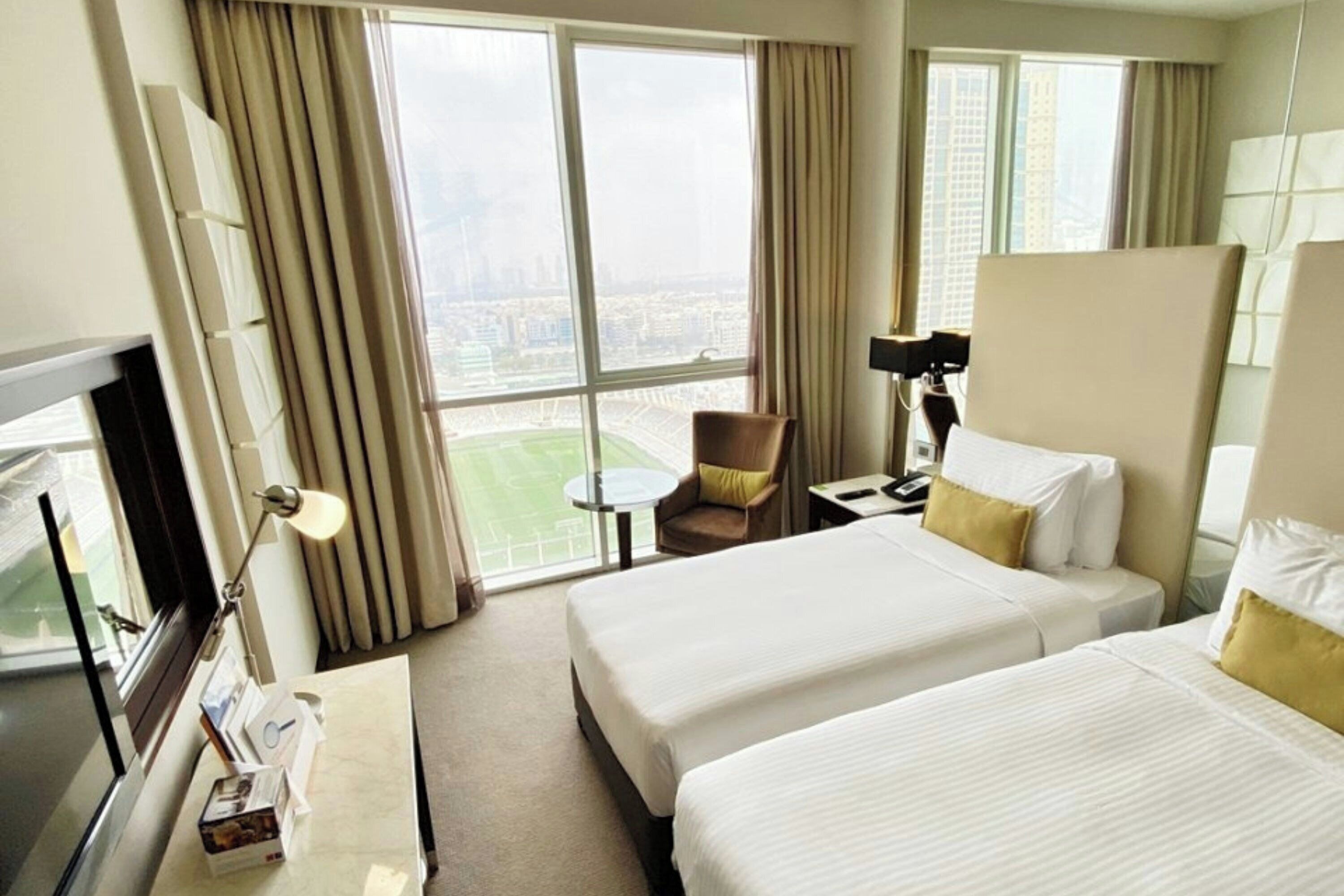 Une chambre d’hôtel avec deux lits, un bureau, une chaise, une télévision et une grande fenêtre donnant sur un paysage urbain.