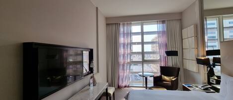 A hotel room with a large bed, a TV, a desk with a chair, a small table, and a view of buildings through the window.