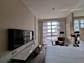 A hotel room with a large bed, a TV, a desk with a chair, a small table, and a view of buildings through the window.