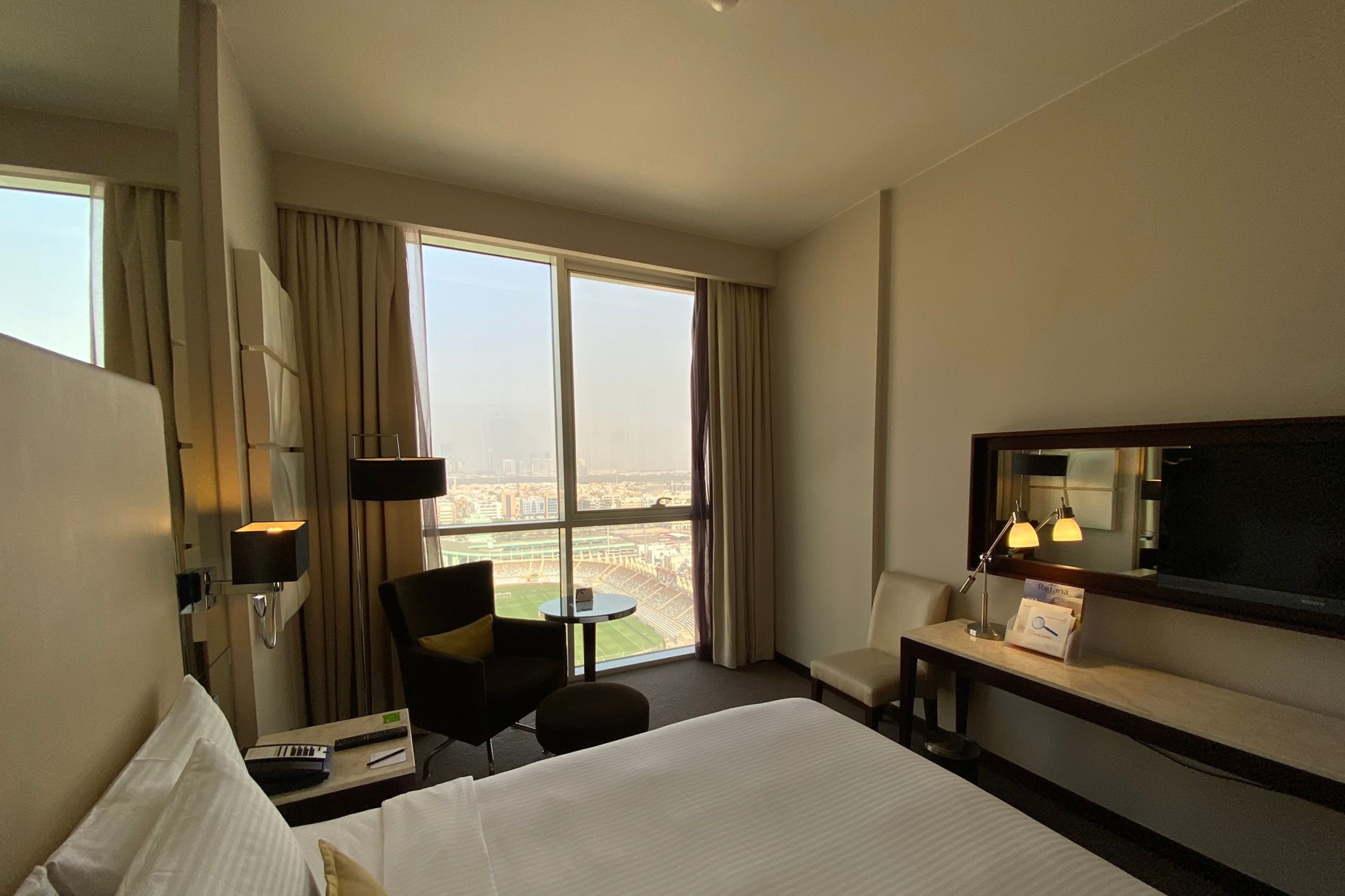 A hotel room with a large bed, a desk, a chair, a TV, and a view of a cityscape through large windows.