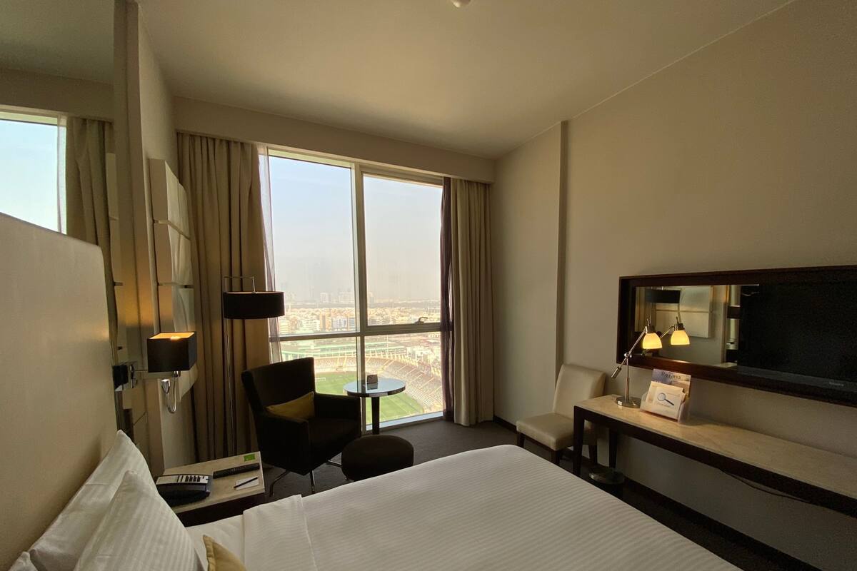 A hotel room with a large bed, a desk, a chair, a TV, and a view of a cityscape through large windows.