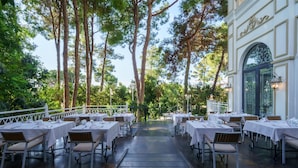 Un restaurante con terraza, mesas y sillas blancas, rodeado de árboles altos.