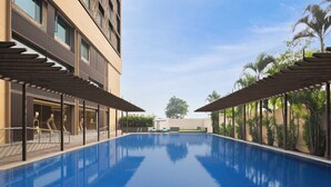 A swimming pool with a clear blue surface, surrounded by a building with large windows and a covered walkway. - Le Meridien Surat (Surat)