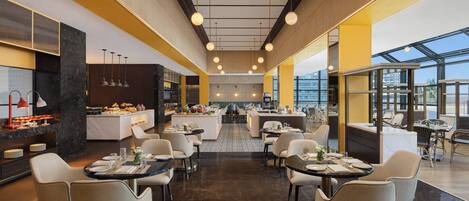 A modern hotel dining area with multiple tables and chairs, a central bar, and large windows offering views.