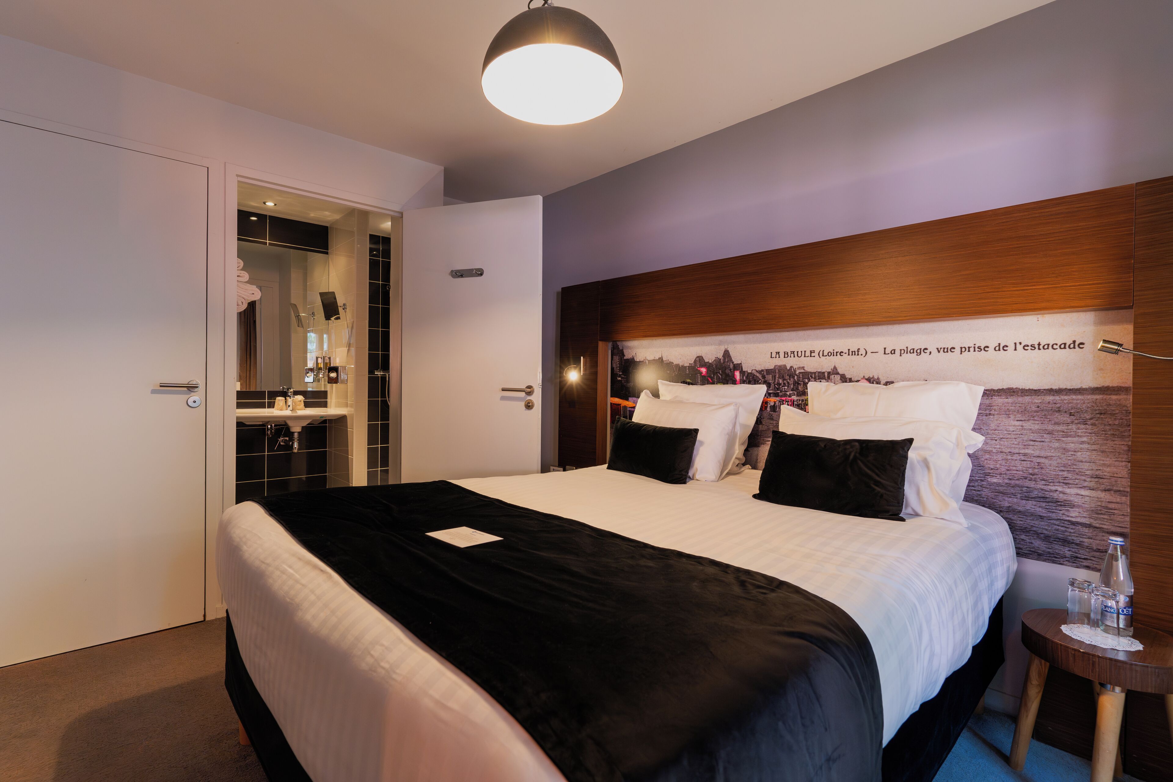 Ein Hotelzimmer mit einem großen Bett, einem Holz-Kopfteil, einem gerahmten Bild an der Wand, einem Nachttisch mit einer Flasche und einem durchgegangenen Tür erkennbaren Badezimmer.