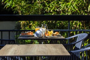 Un tavolo con prodotti per la colazione, un bollitore e un bicchiere di succo d'arancia su un balcone esterno con ringhiere in metallo e vegetazione sullo sfondo.