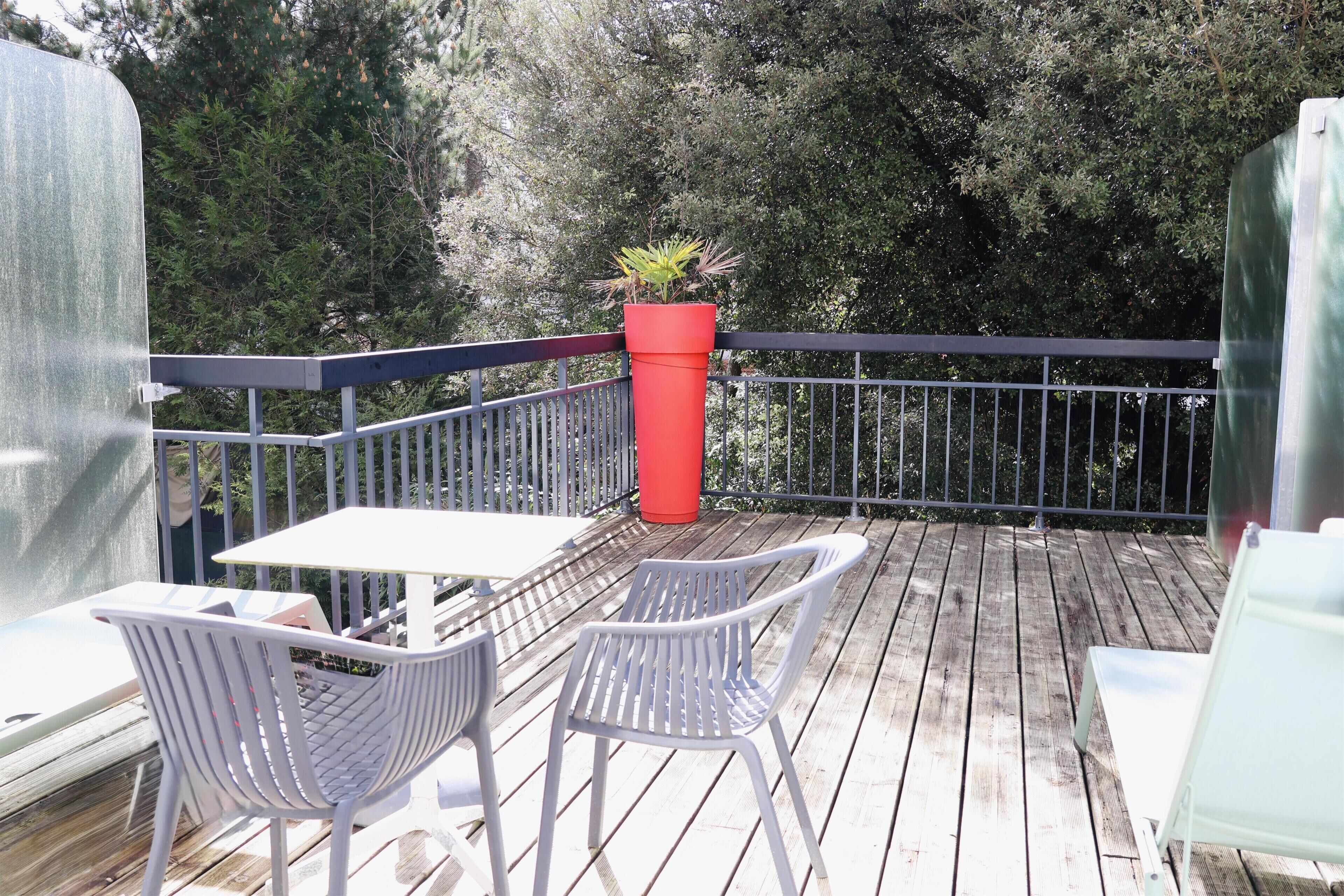 Una terraza de madera con una mesa y dos sillas, una maceta roja con plantas y una barandilla metálica.