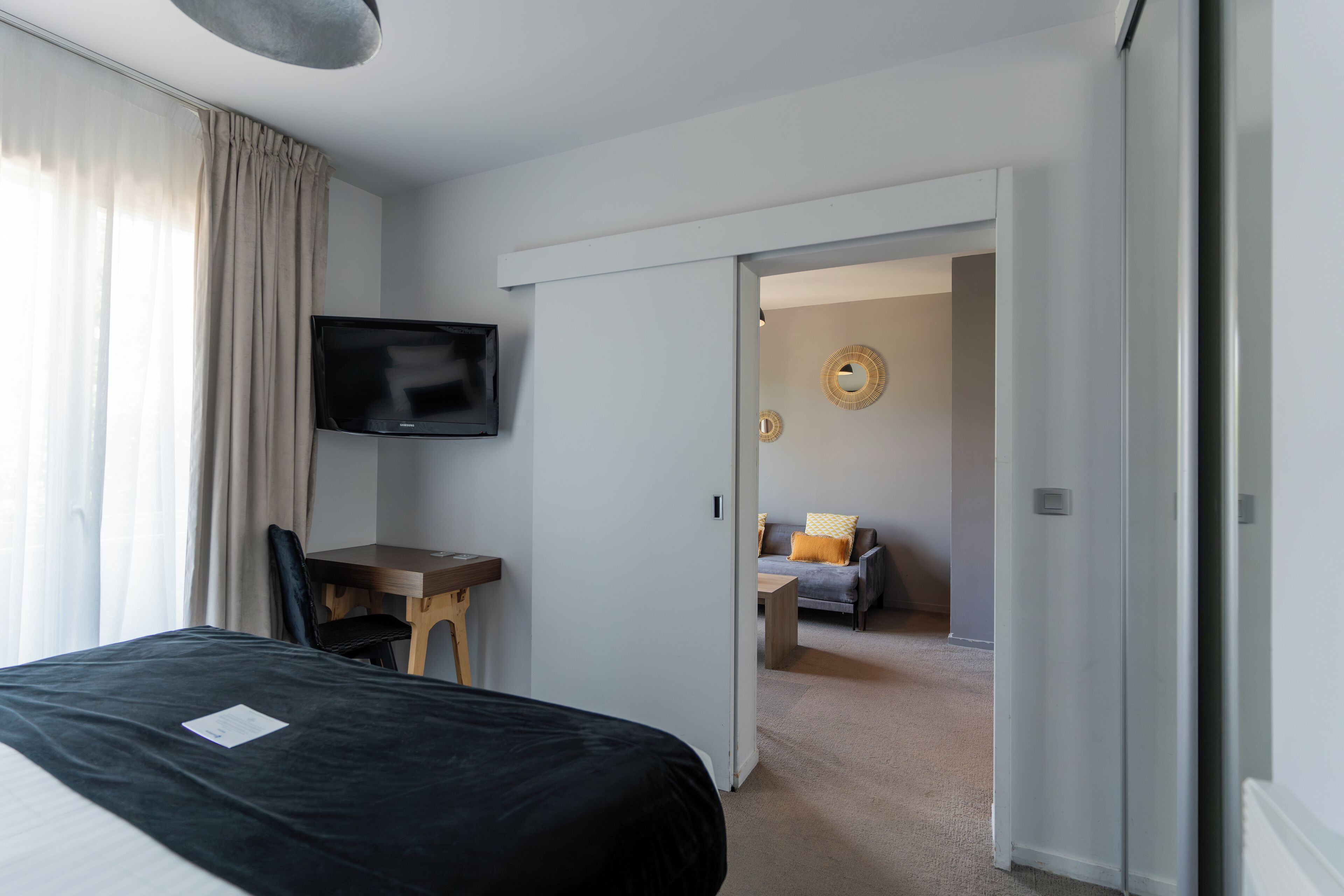 Habitación de hotel con cama, escritorio con silla, televisor montado en la pared y vista a un área de estar contigua.