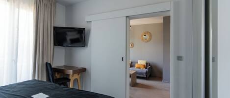 Habitación de hotel con cama, escritorio con silla, televisor montado en la pared y vista a un área de estar contigua.