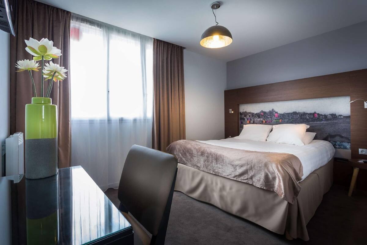 A hotel room with a bed, a desk, a chair, a vase with flowers, a cityscape painting, and a pendant light.