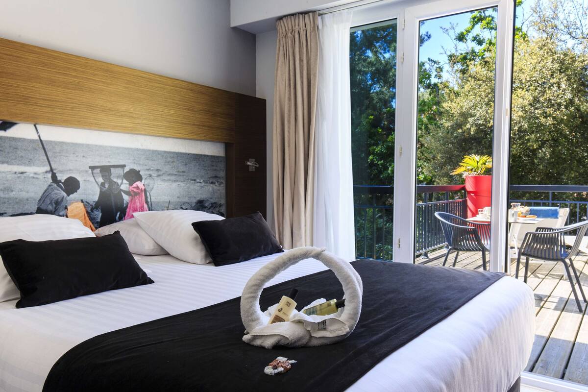 A hotel room with a bed, a painting on the wall, a balcony with chairs and a table, and a window with curtains.