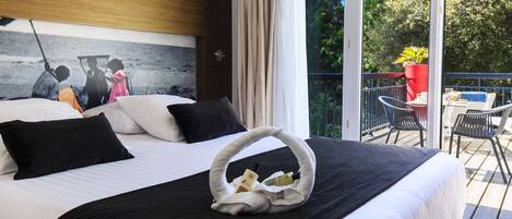 Quarto de hotel com cama, um quadro na parede, varanda com cadeiras e uma mesa, e janela com cortinas.
