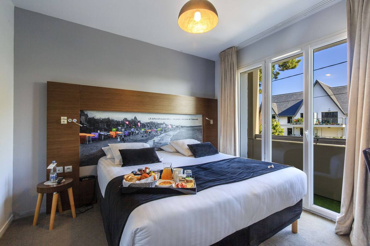 A hotel room with a large bed, a cityscape poster on the wall, a tray with breakfast items, and a view of the outside balcony.