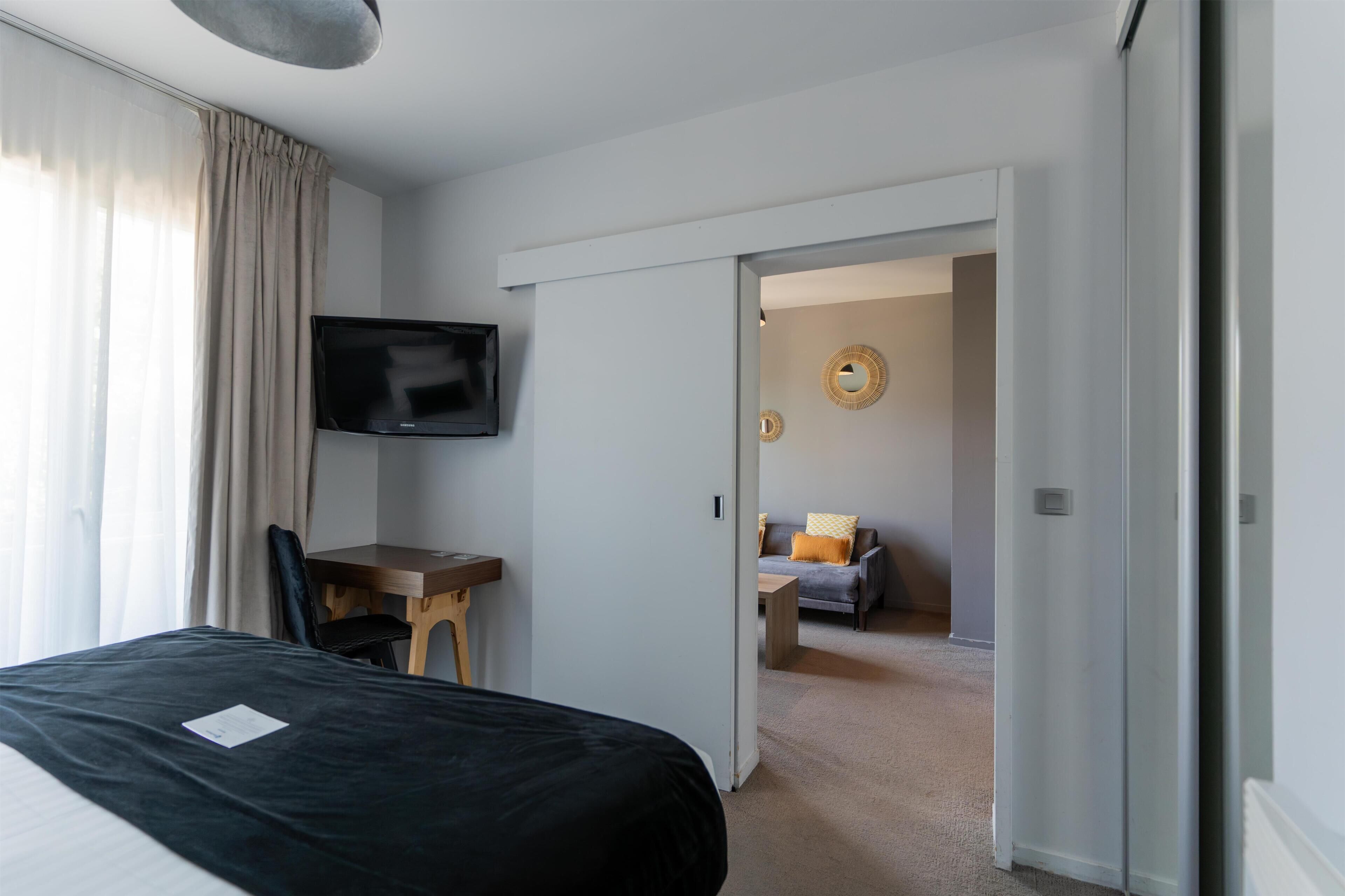 Habitación de hotel con cama, escritorio con silla, televisor y vista a un área de estar contigua.