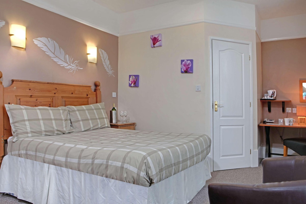A bedroom with a wooden headboard, a bed with a plaid bedspread, a nightstand with a bottle and glasses, and wall-mounted light fixtures.