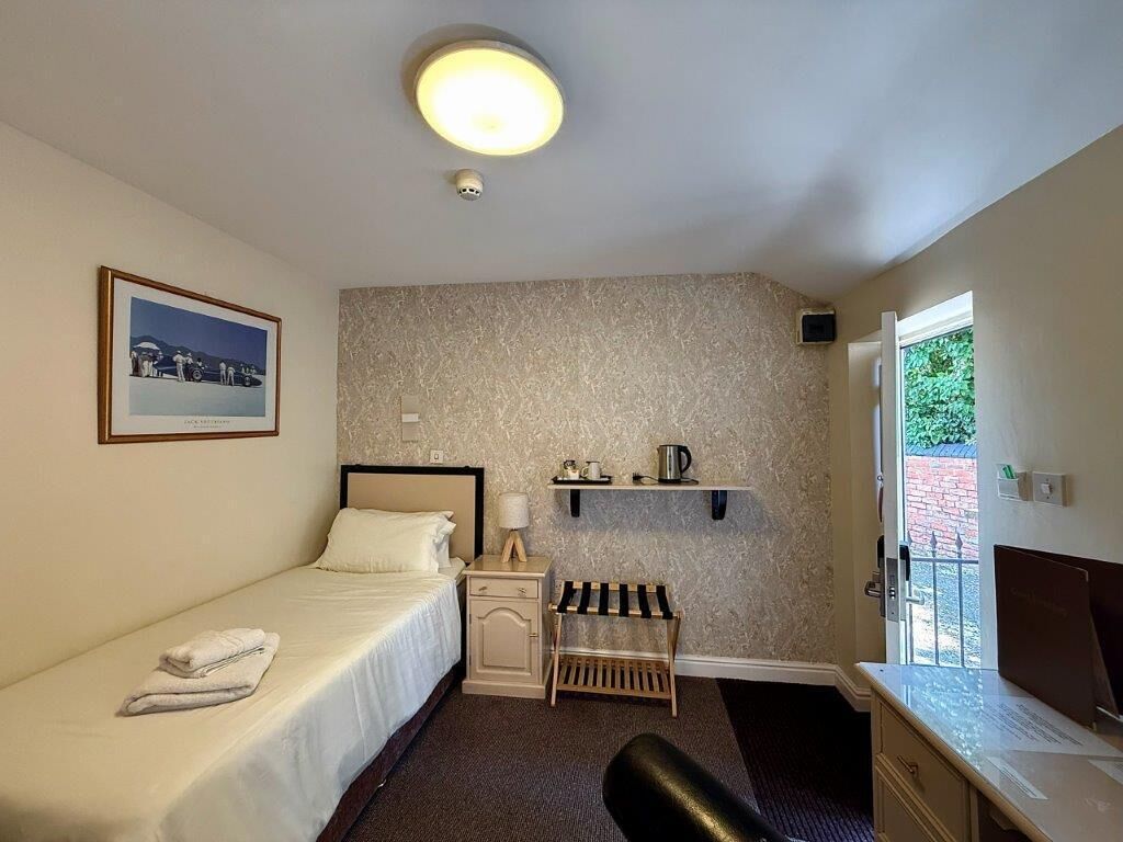 A small, neatly arranged room with a bed, bedside table, lamp, and a shelf with a kettle.
