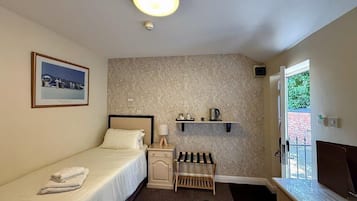 A small, neatly arranged room with a bed, bedside table, lamp, and a shelf with a kettle.