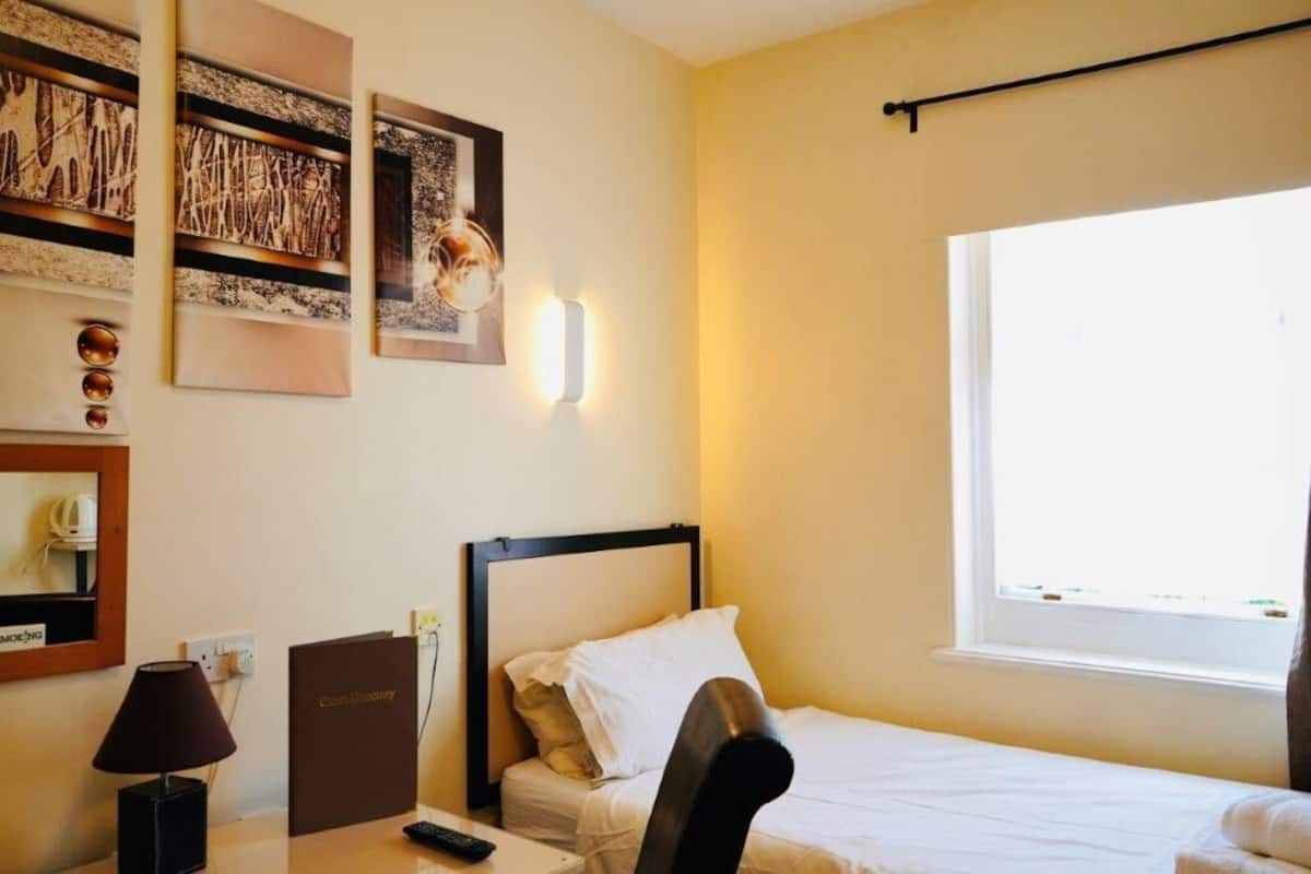 A hotel room with a bed, a desk with a lamp, a chair, a bedside table, a bedside lamp, a wall-mounted light, a window, and artwork on the wall.