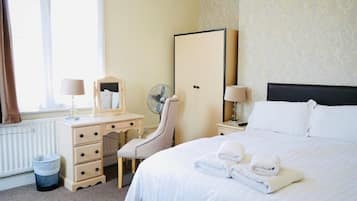 Habitación de hotel con cama, escritorio con cajones, silla, espejo, mesita de noche con lámpara y radiador.