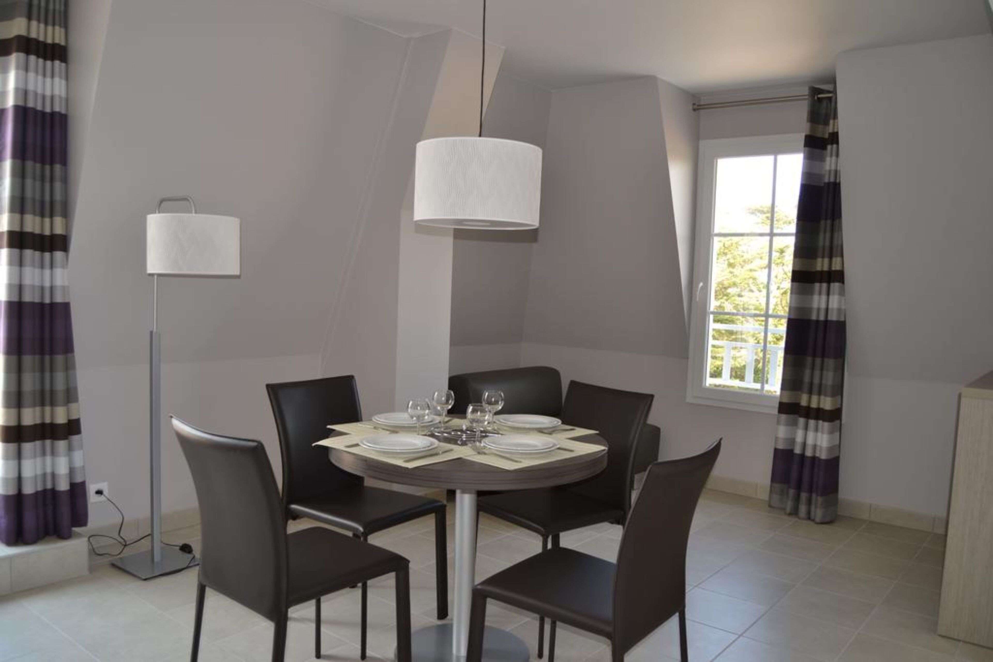 A dining area with a round table set for four, black chairs, a hanging light fixture, and a window with curtains.