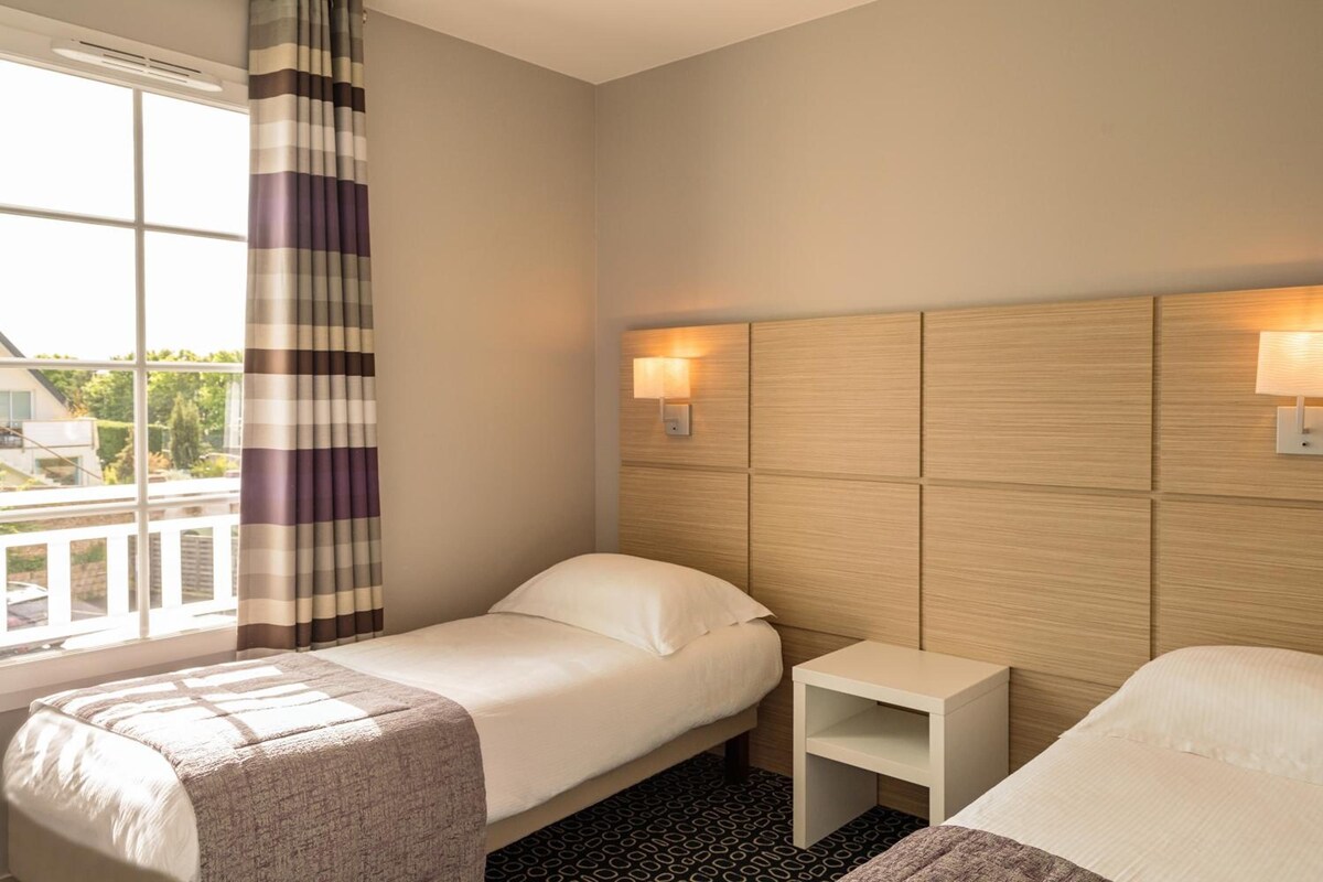A hotel room with two beds, a window with curtains, a bedside table, and a headboard with wall-mounted lights.