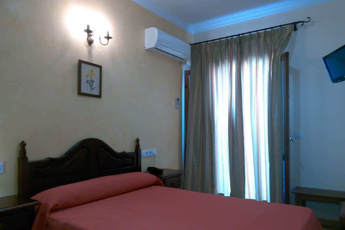 A bedroom with a bed, a nightstand, a wall-mounted air conditioner, a framed picture, and a curtain.