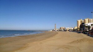 A wide sandy beach with a clear blue sky, a tall lighthouse, and buildings along the coastline.