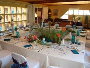 Uma sala de jantar com mesas preparadas para uma refeição, com copos de vidro azul e arranjos florais.