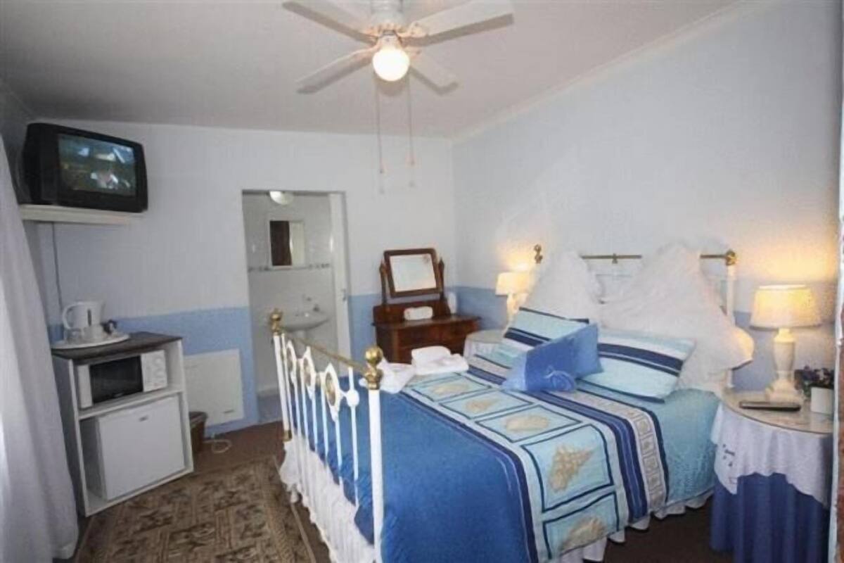 Habitación de hotel con cama, mesitas de noche, televisor, ventilador de techo y una pequeña cocina americana.
