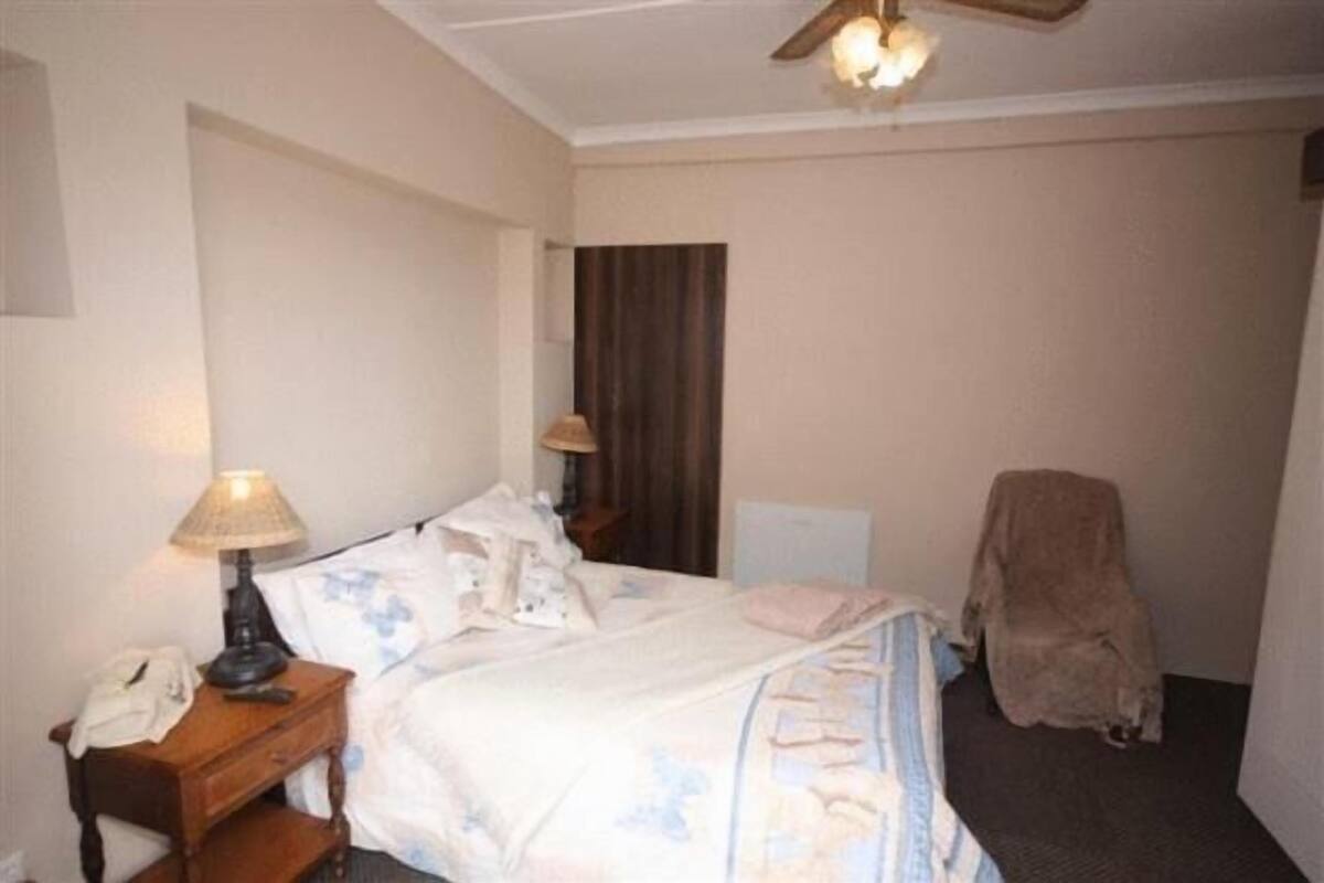 Habitación de hotel con cama, mesita de noche con lámpara, silla y perchero.