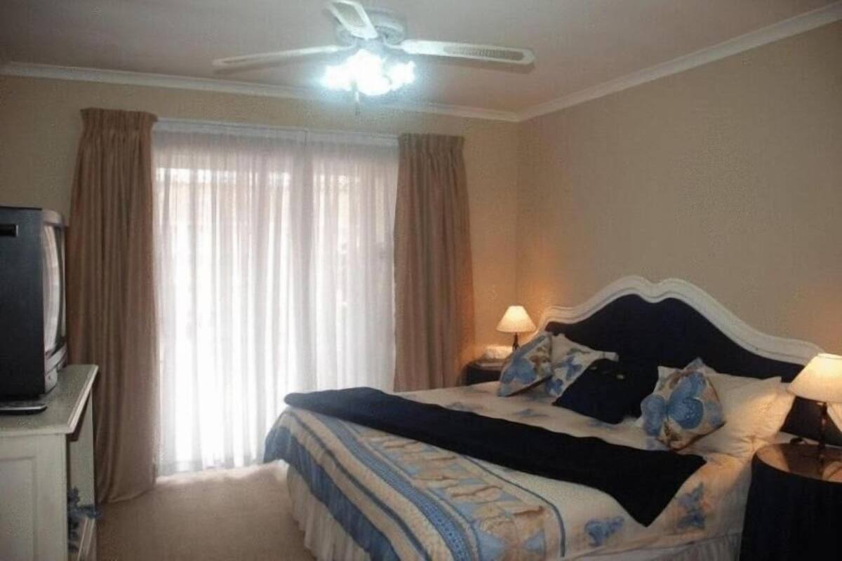 Un dormitorio con cama, cortinas, ventilador de techo, lámpara y televisor sobre un mueble.
